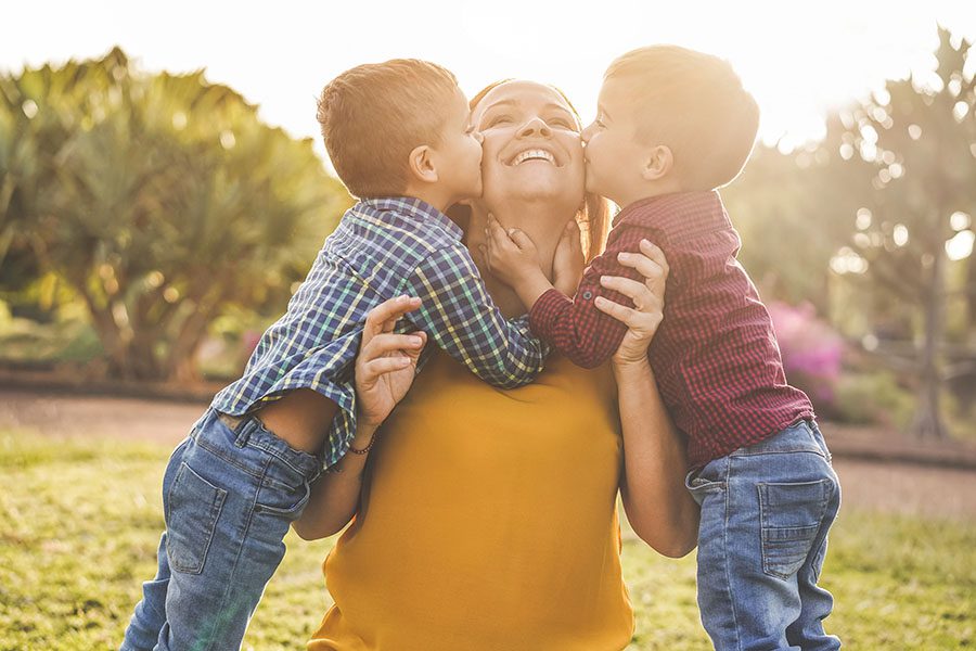 About Our Agency - Happy Mother Being Kissed on the Cheeks by Her Two Sons in the Backyard